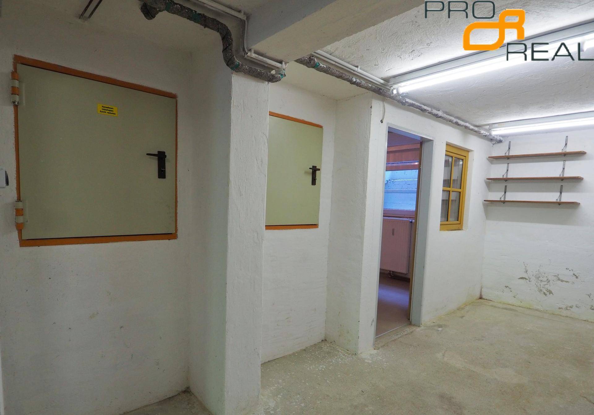 Kellerraum mit Betonboden, sichtbaren Leitungen und einfachen Lagertüren.