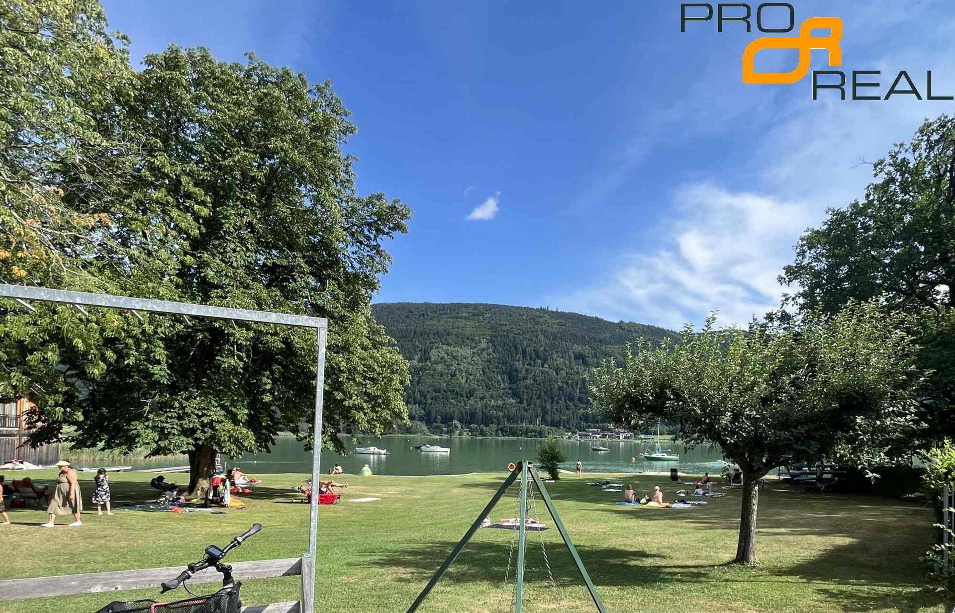 Badestrand am See mit Liegewiese, Bäumen und beeindruckender Bergkulisse im Hintergrund.