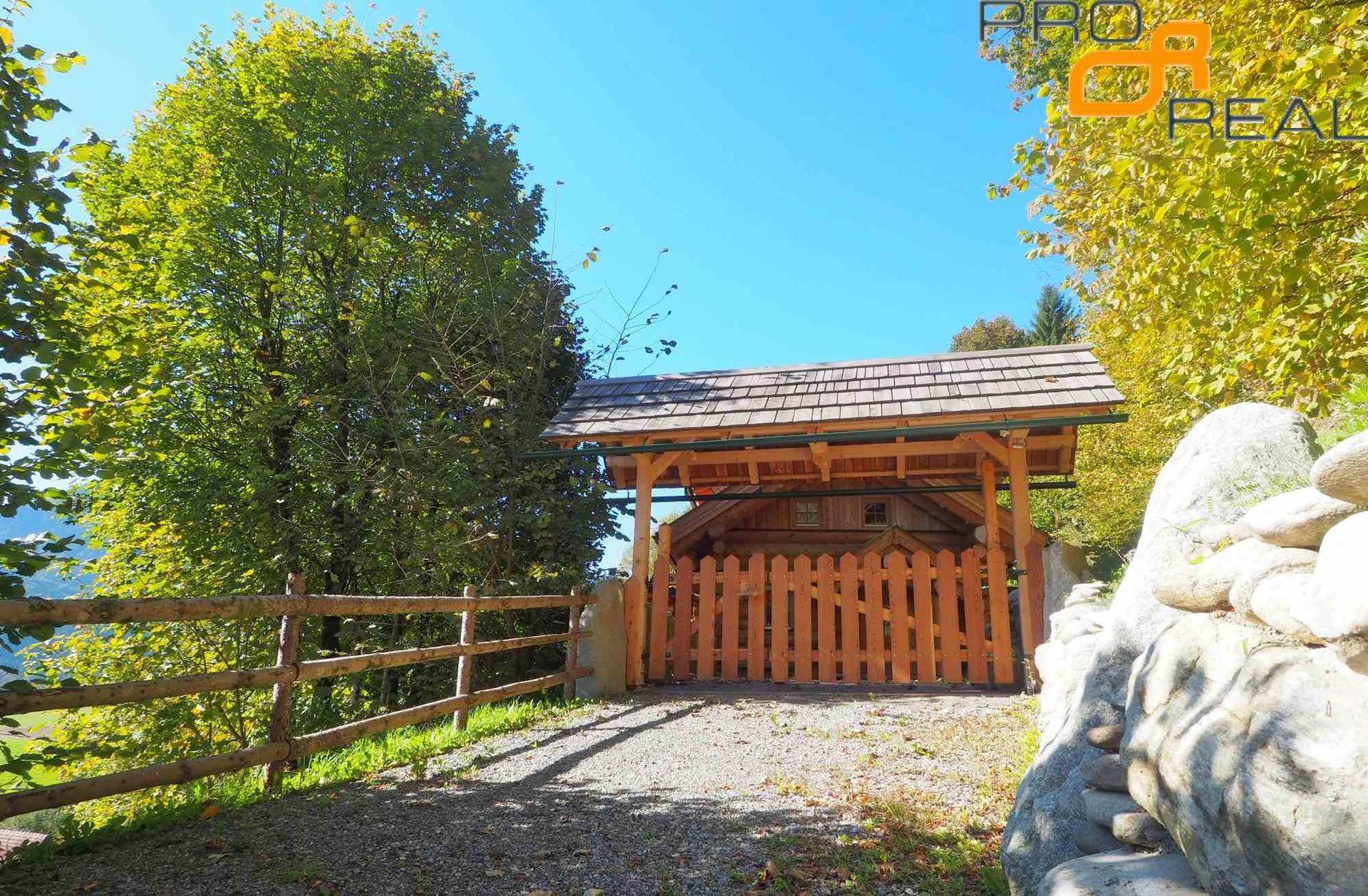 Hölzerner Carport mit Zaun und Schindeldach, umgeben von Bäumen.