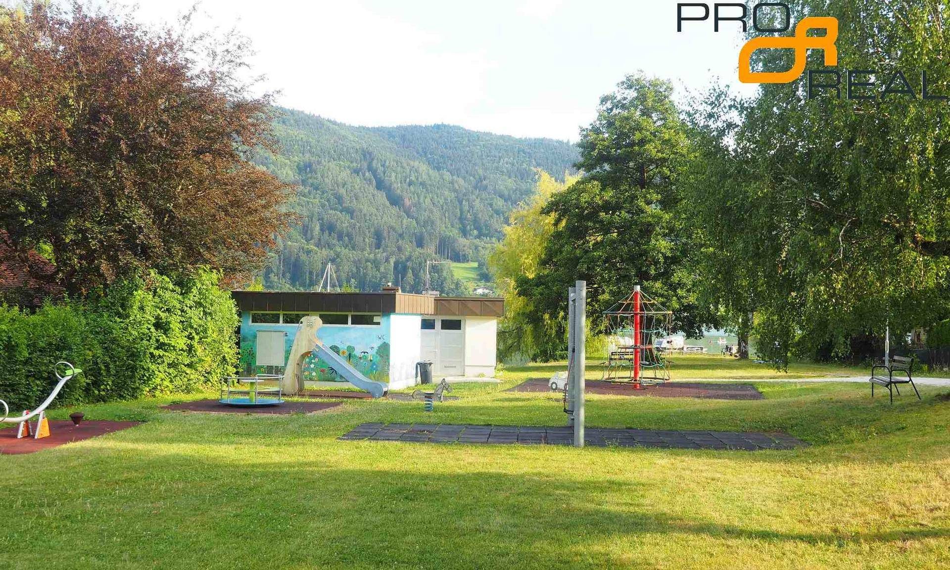 Kinderspielplatz mit Rutsche, Wippe und Karussell, umgeben von einer grünen Wiese und Bäumen.