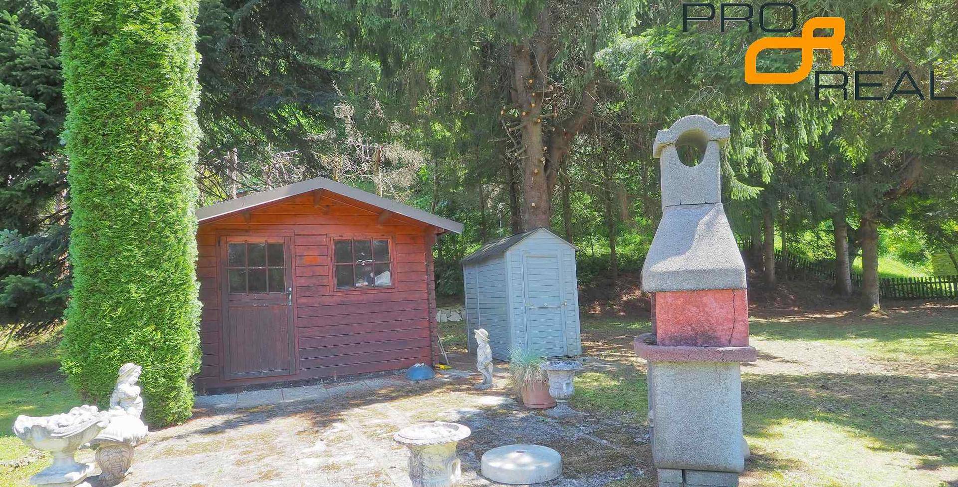 Gartenbereich mit zwei Gartenhäusern, einem Grillplatz und hohen Bäumen im Hintergrund.