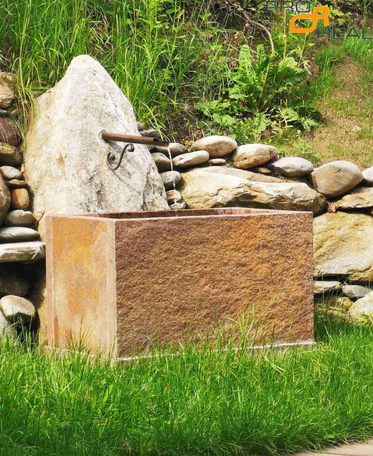 Rustikaler Steinbrunnen im Garten, umgeben von Felsen und Grün.