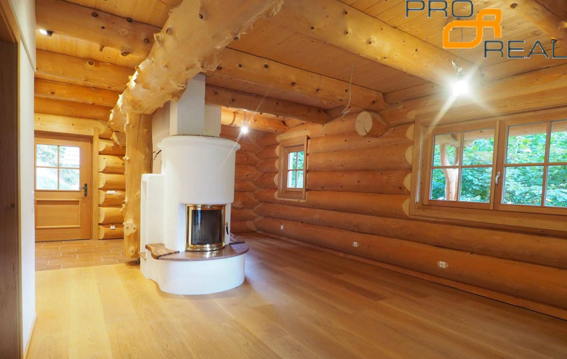Heller Wohnraum mit Holzbalken, Kamin und Blick ins Grüne durch die Fenster.