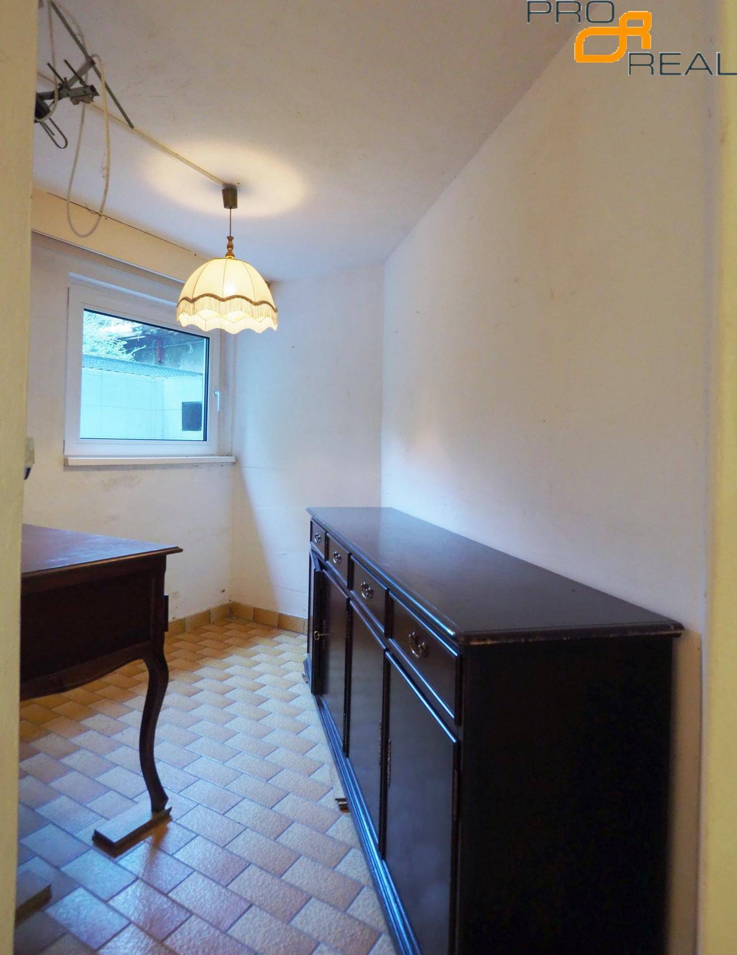 Kellerraum mit Fliesenboden, Fenster und einem dunklen Holzschrank, der als Stauraum dient.