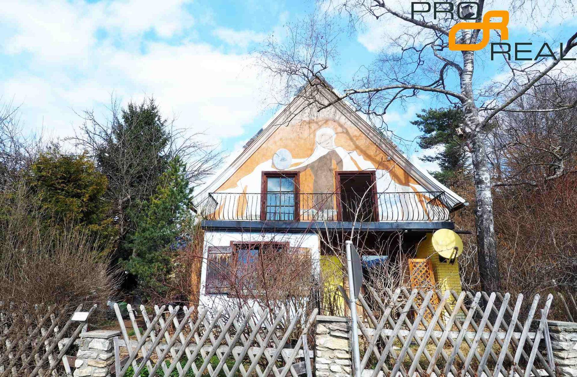 Giebelansicht des Hauses mit Balkon, umgeben von Bäumen und einem Holzzaun im Vordergrund.