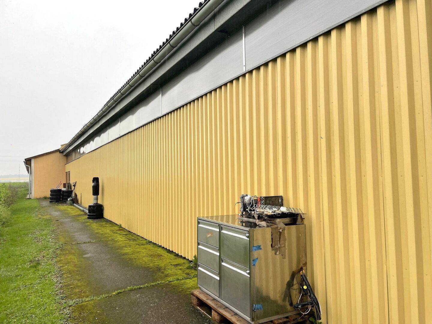 Lange gelbe Wellblechfassade des Gebäudes mit einem schmalen Pfad und Grasstreifen daneben.