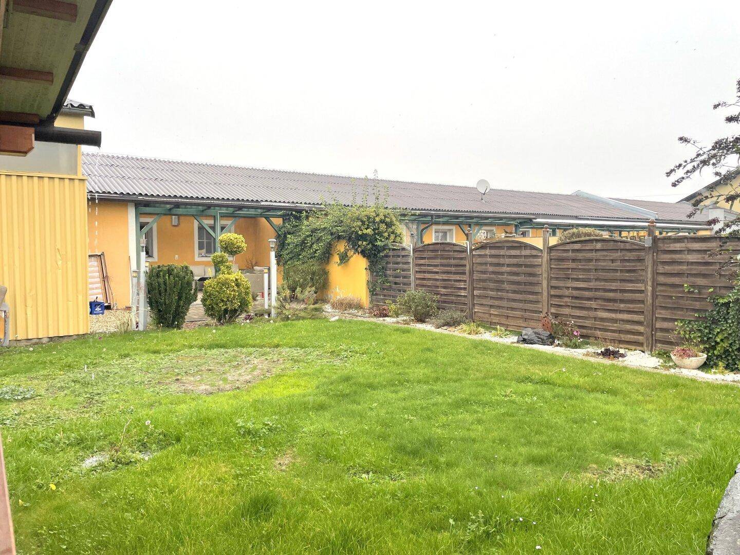 Gepflegter Gartenbereich mit Rasenfläche, Holzzaun und einem überdachten Sitzbereich im Hintergrund.