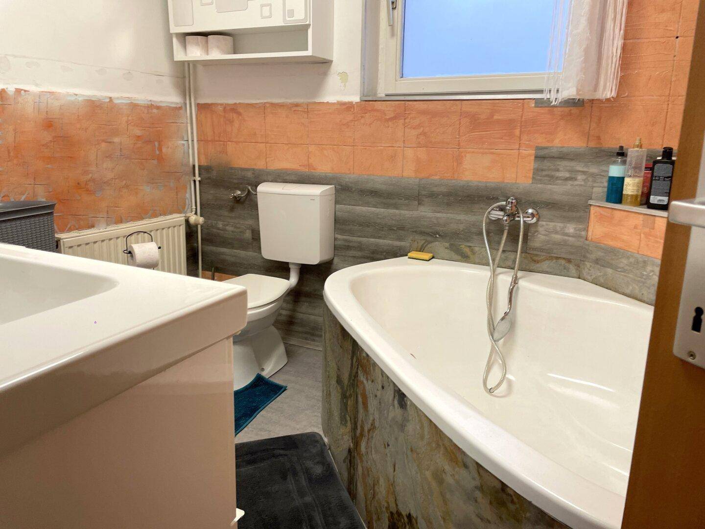 Badezimmer mit Badewanne, Toilette und teilweise gefliesten Wänden in Orange- und Grautönen.