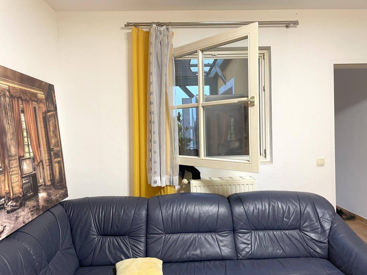 Wohnzimmer mit blauem Sofa und einem geöffneten Fenster, das viel Tageslicht hereinlässt.