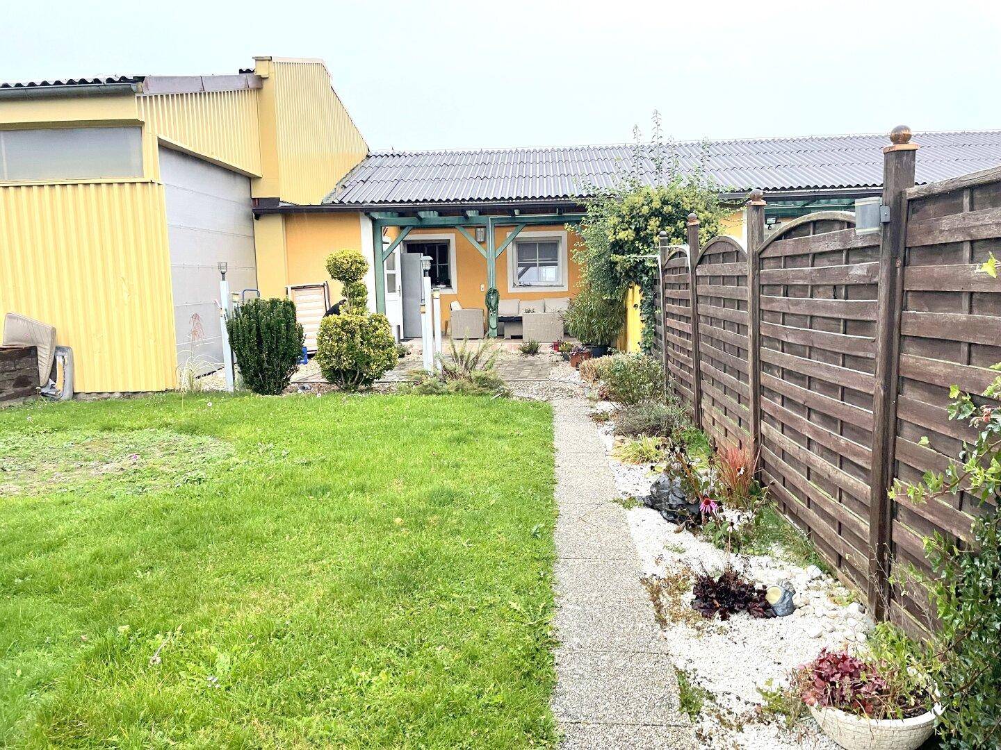 Garten mit Rasenweg, Blumenbeeten und Blick auf die Rückseite des Hauses mit überdachter Terrasse.