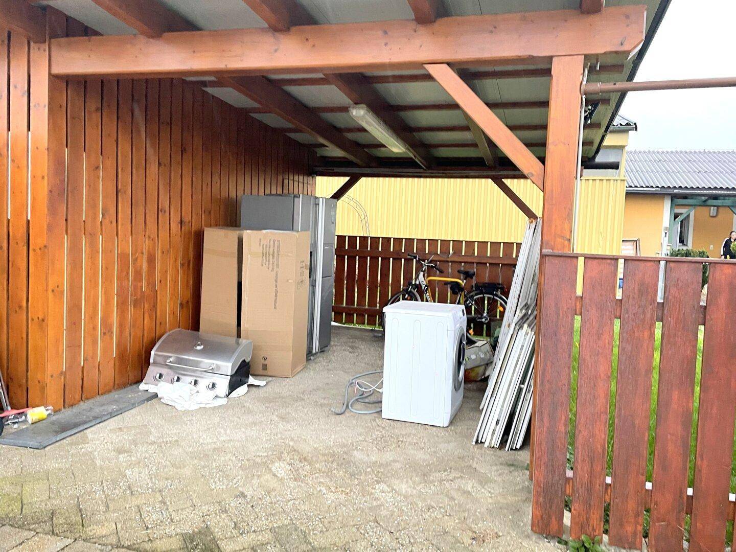Überdachter Holzunterstand mit Waschmaschine, Fahrrädern und Grillgeräten.