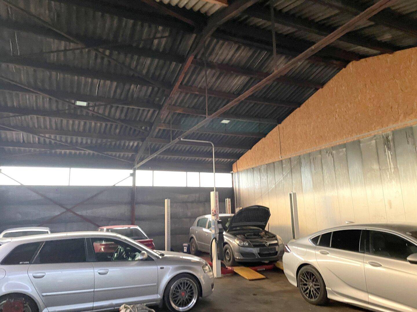 Innenansicht einer großen Werkstatt mit Autos, Hebebühnen und sichtbarer Dachkonstruktion.