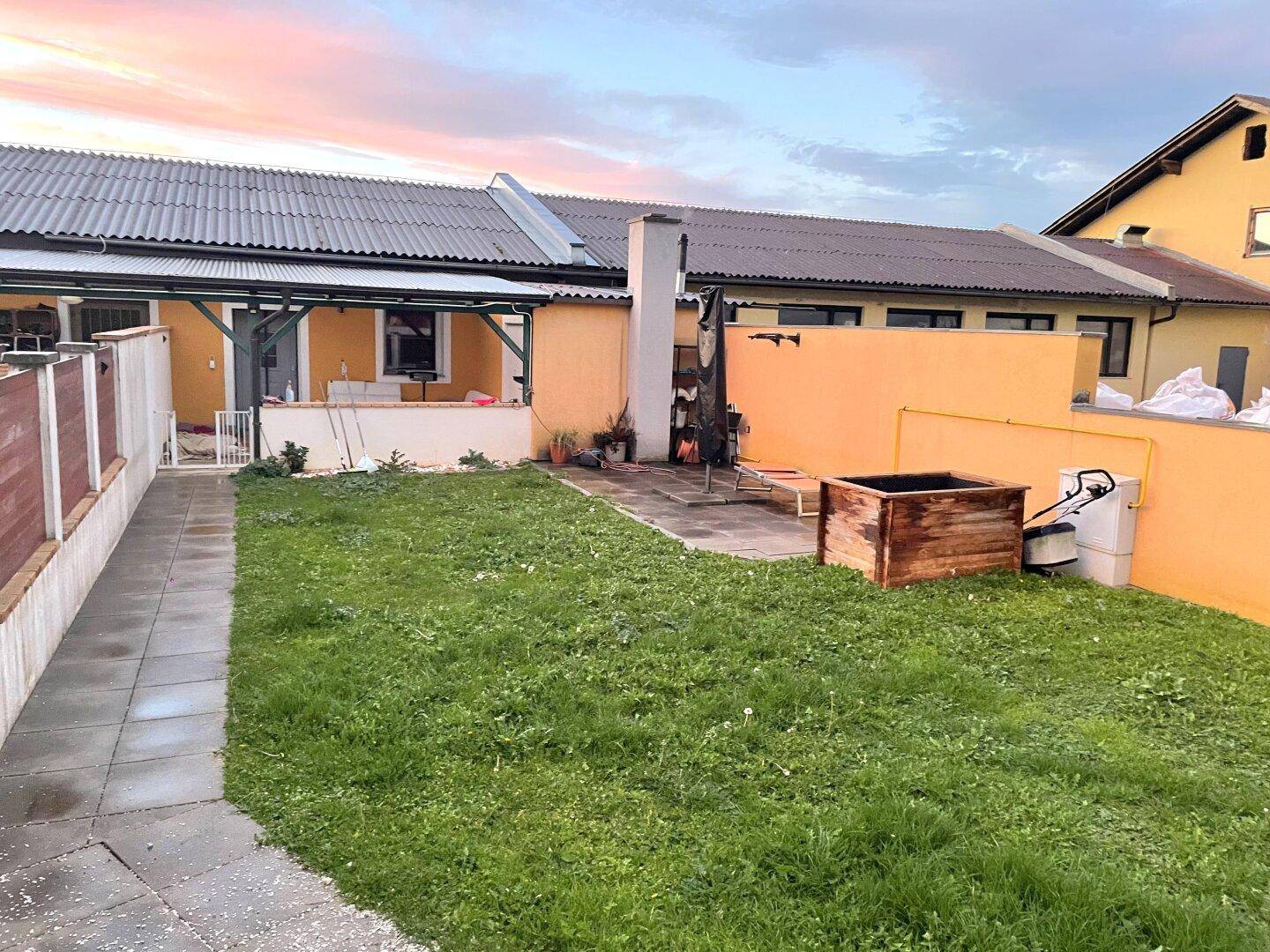 Gartenbereich am Abend mit Rasenfläche, gepflastertem Sitzplatz und einem Grillkamin.