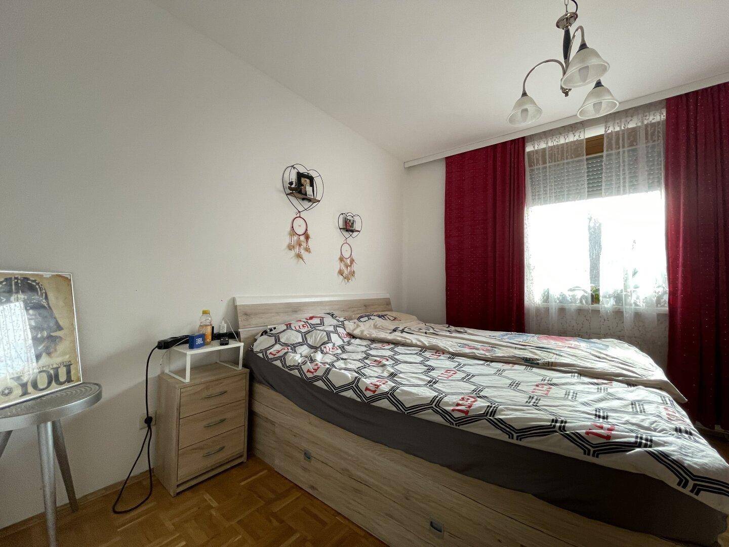 Gemütliches Schlafzimmer mit Doppelbett, Parkettboden und roten Vorhängen am Fenster.