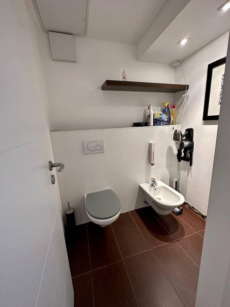 Separate Toilette mit Bidet, Wand-WC und braunem Fliesenboden.