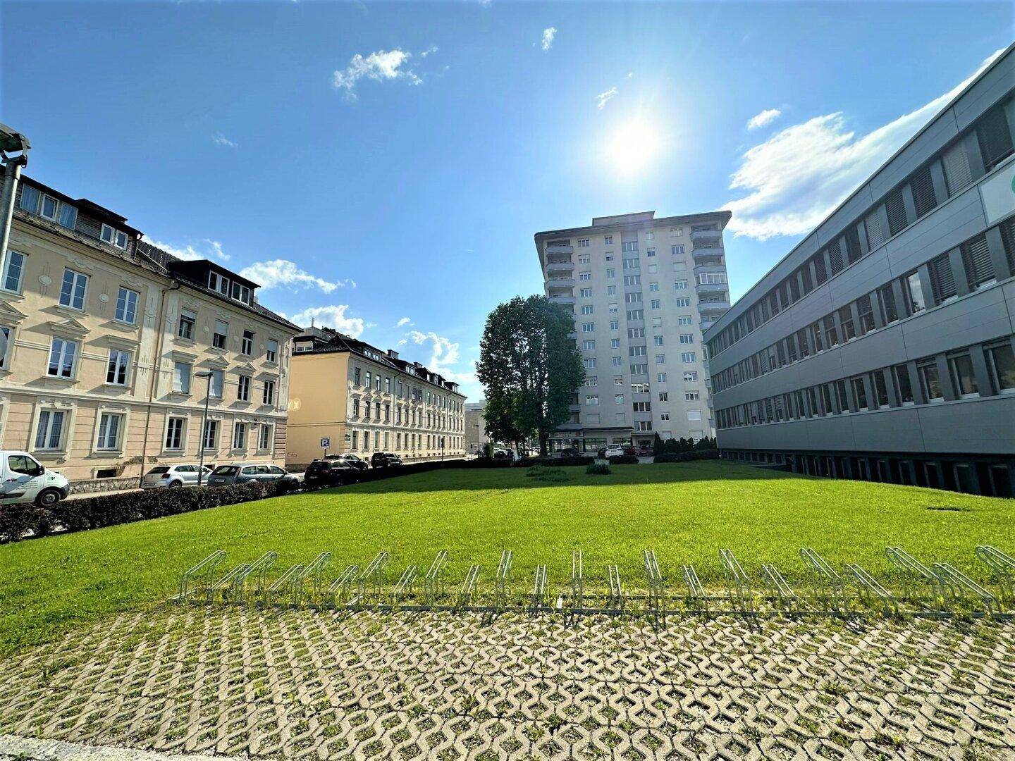 Grüne Rasenfläche vor modernen und klassischen Gebäuden unter strahlendem Sonnenschein.