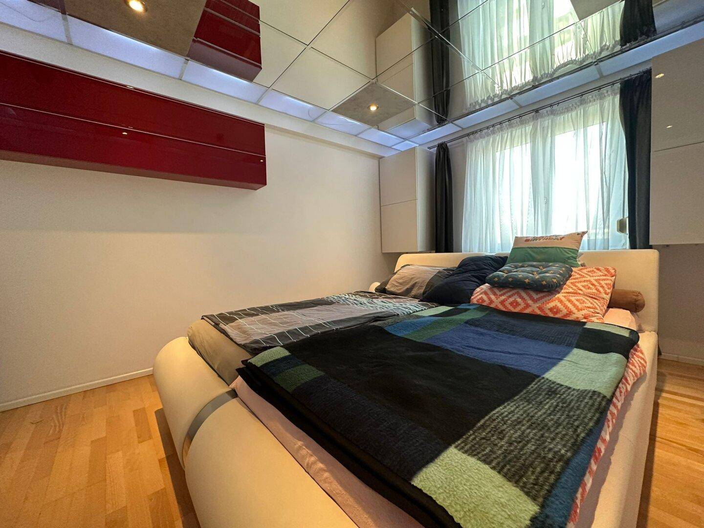 Schlafzimmer mit modernem Doppelbett, Parkettboden und großen Fenstern mit Vorhängen.