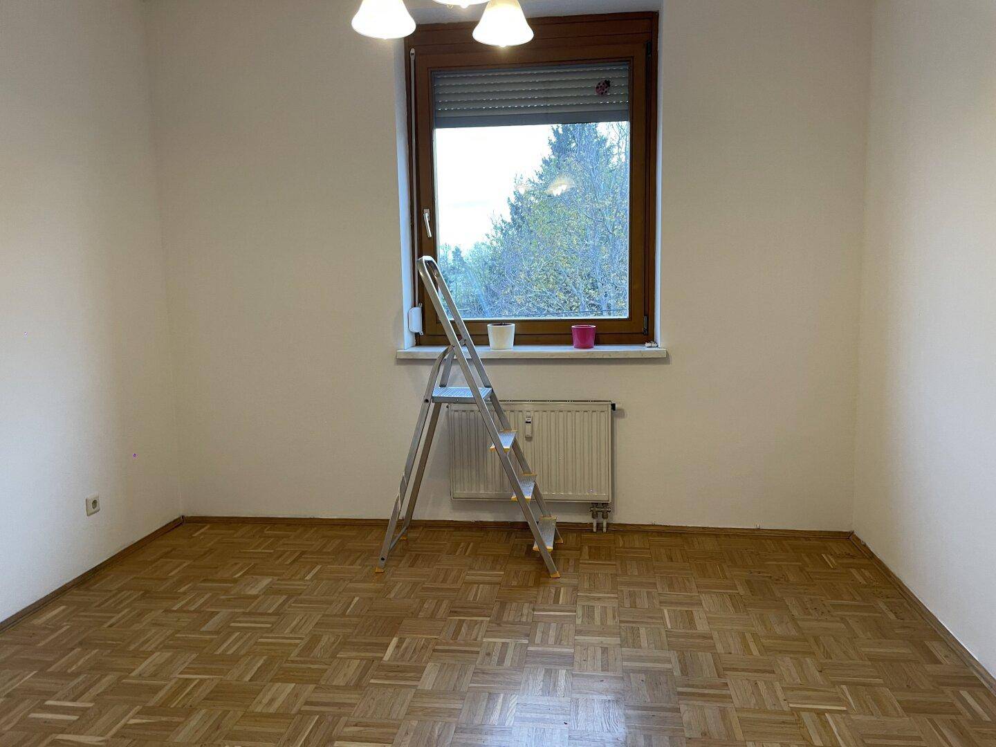 Leeres Zimmer mit Parkettboden und einem Fenster, das einen Blick ins Grüne bietet.
