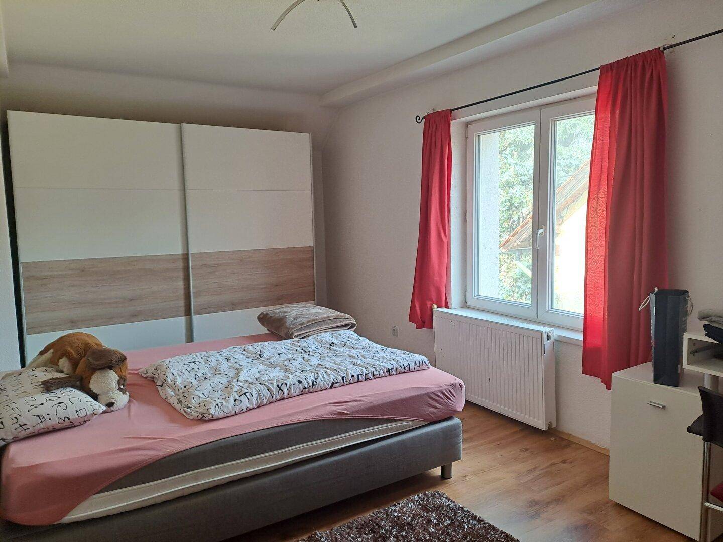 Schlafzimmer mit Doppelbett, großem Schrank und Fenster mit rotem Vorhang.