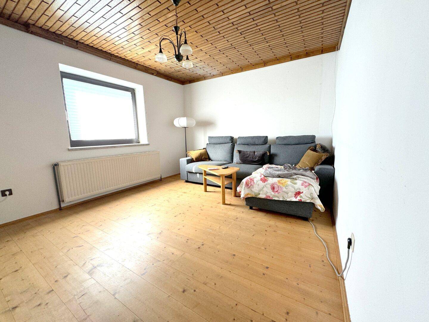 Helles Wohnzimmer mit Holzbalkendecke, Holzboden und einem grauen Sofa.