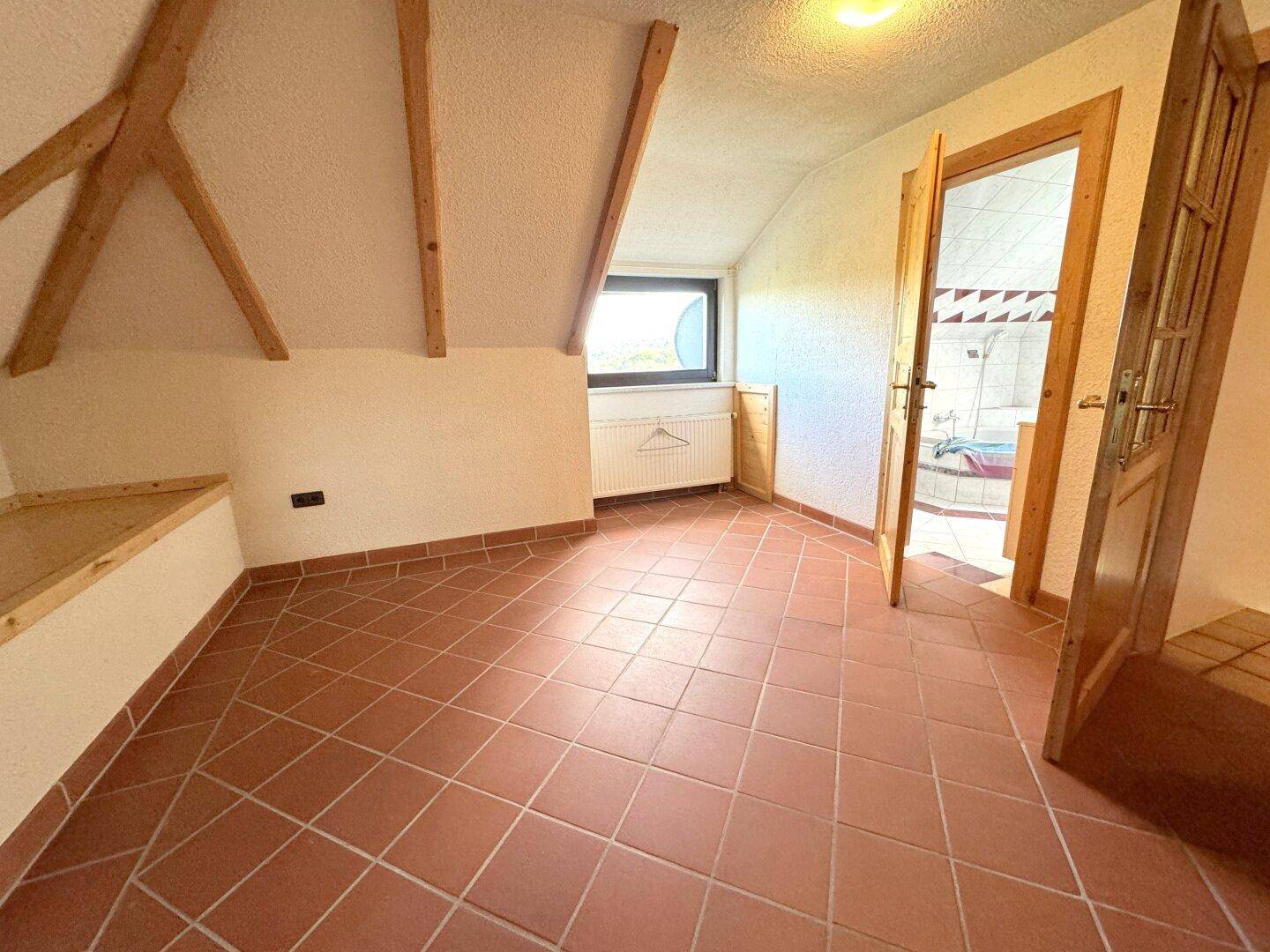 Dachgeschossraum mit Terrakottafliesen, Holzbalken und Zugang zum Badezimmer.