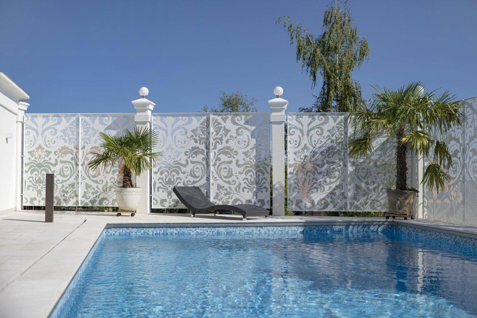 Detailansicht des Pools mit Liegestuhl und dekorativer Zaunwand, umgeben von Palmen.