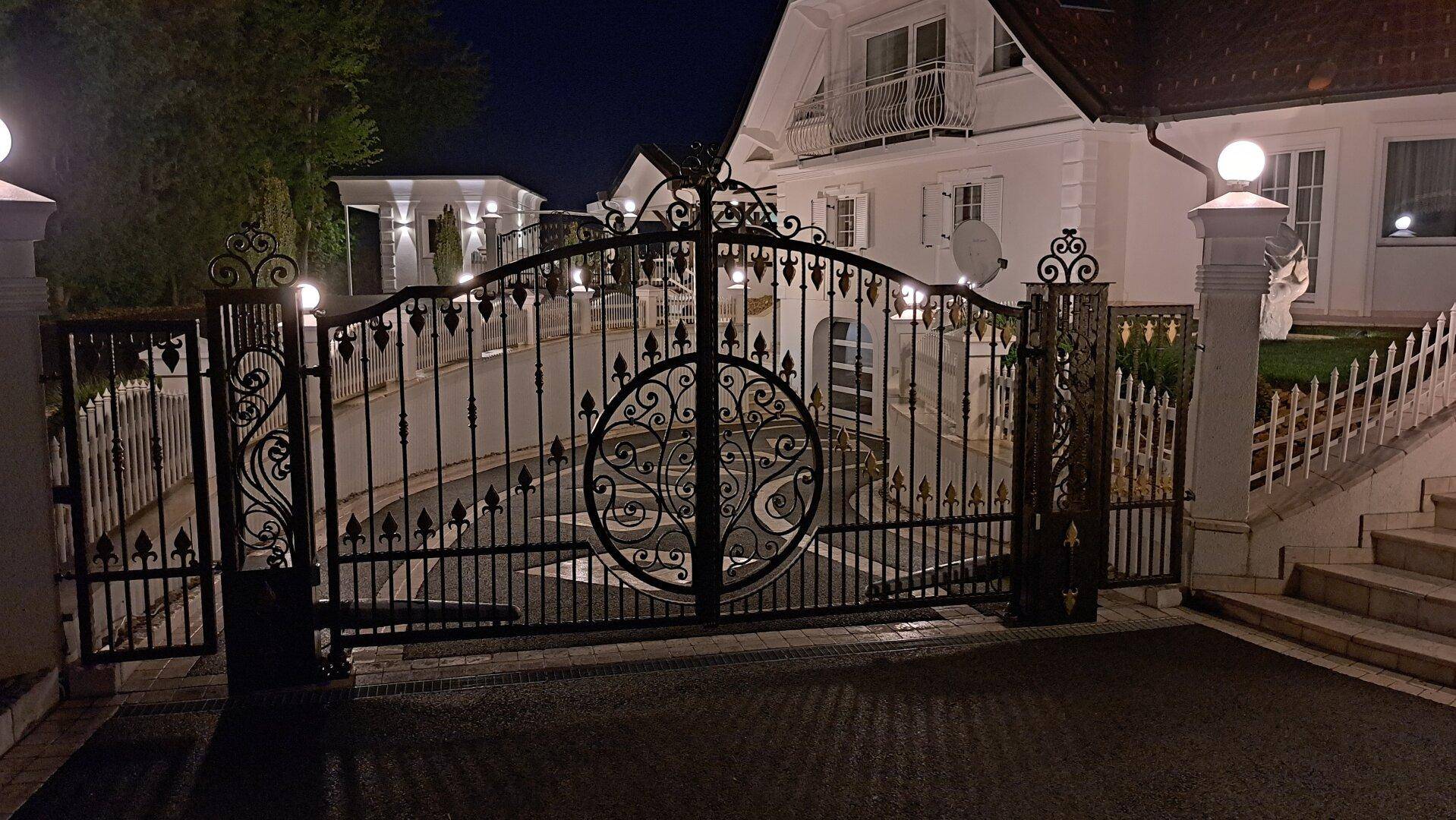 Detailansicht des schmiedeeisernen Eingangstors bei Nacht, kunstvoll verziert und beleuchtet.
