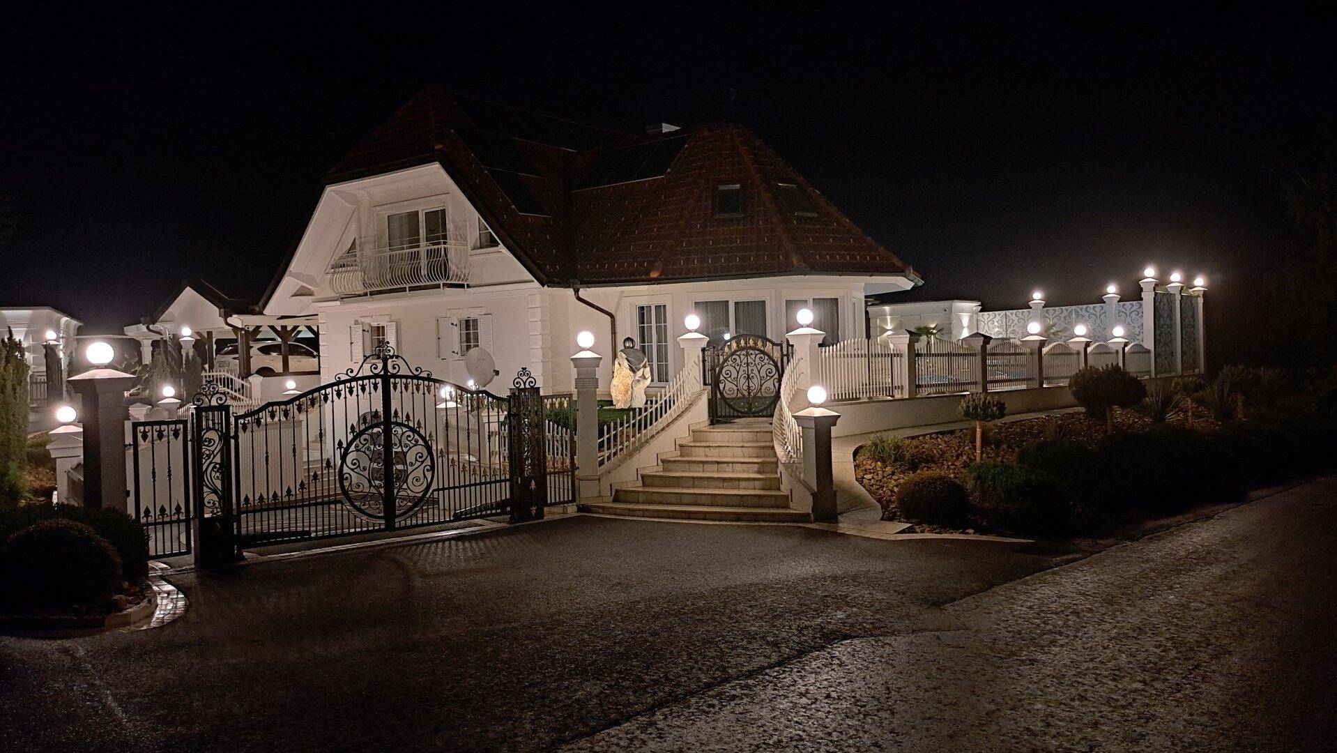 Das Haus bei Nacht, hell beleuchtet, mit schmiedeeisernem Tor und gepflegter Auffahrt.