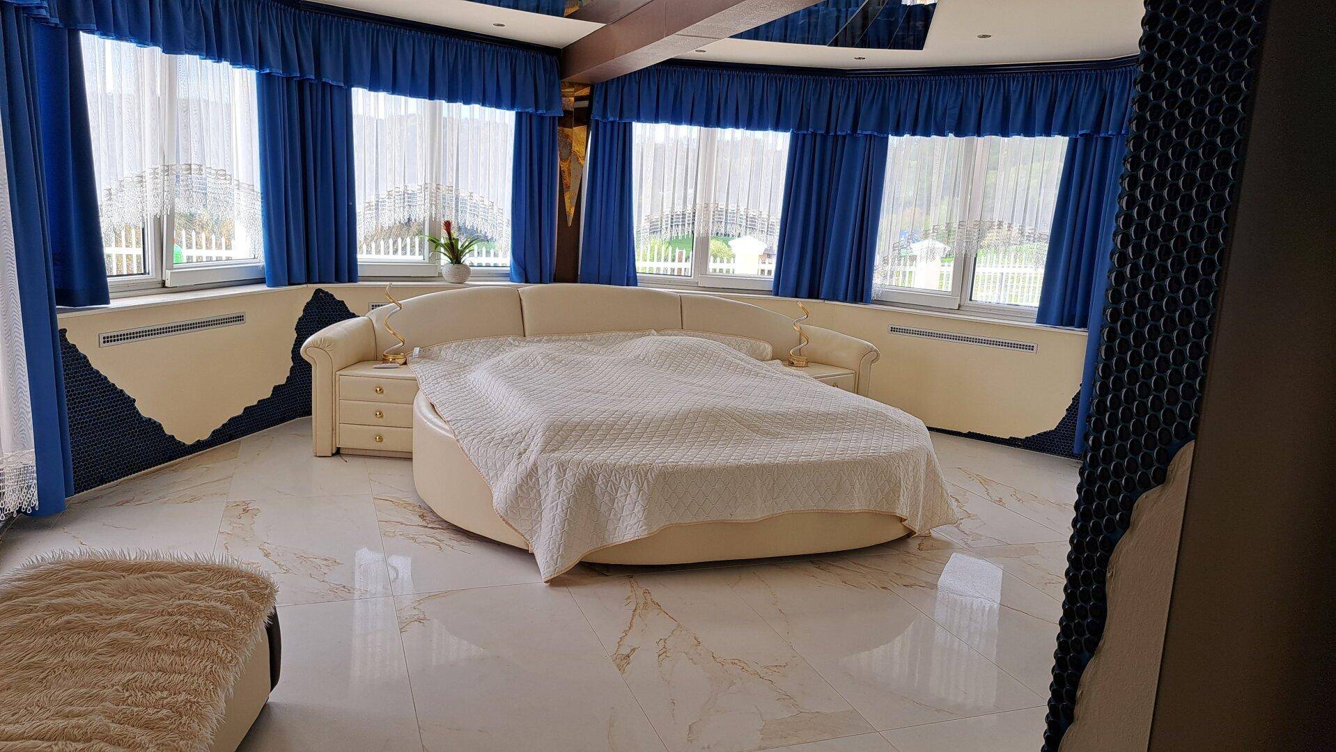 Großes Schlafzimmer mit rundem Bett, Marmorboden und vielen Fenstern mit blauen Vorhängen.