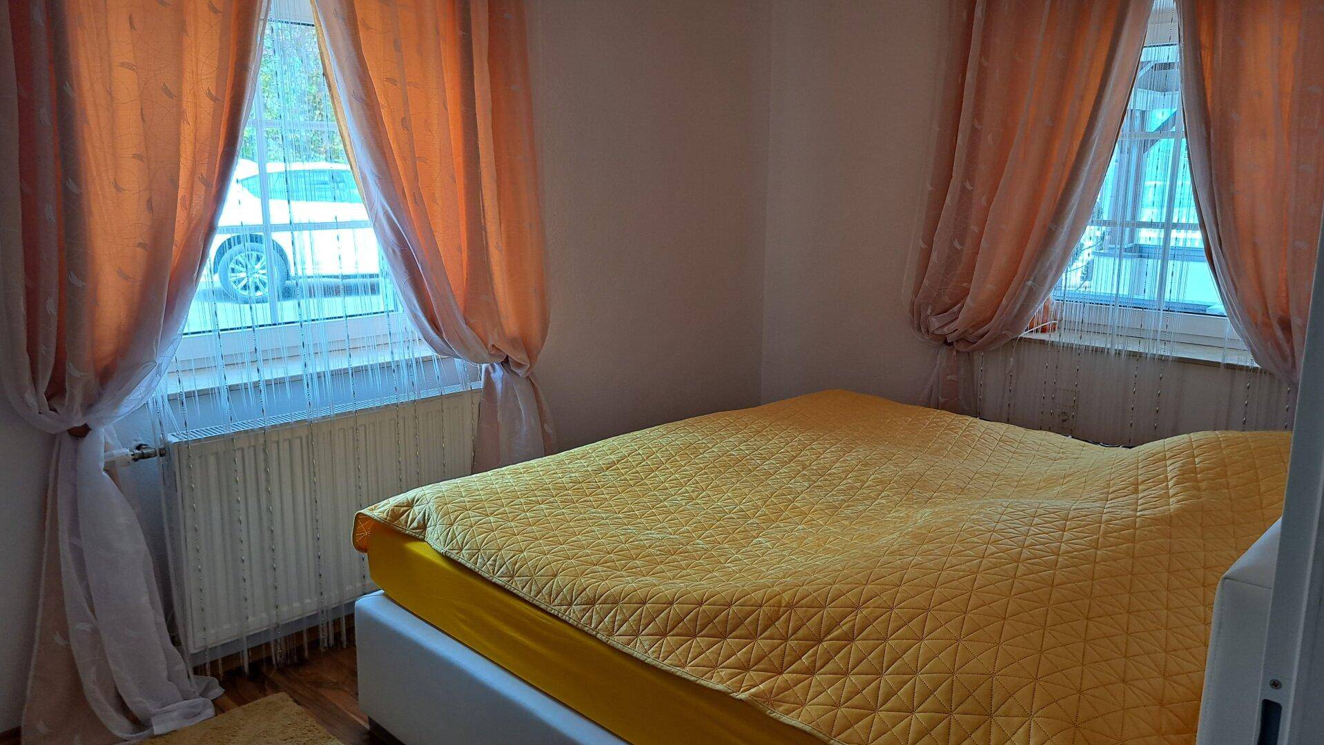Kleines Schlafzimmer mit gelber Bettwäsche und zwei Fenstern mit orangefarbenen Vorhängen.