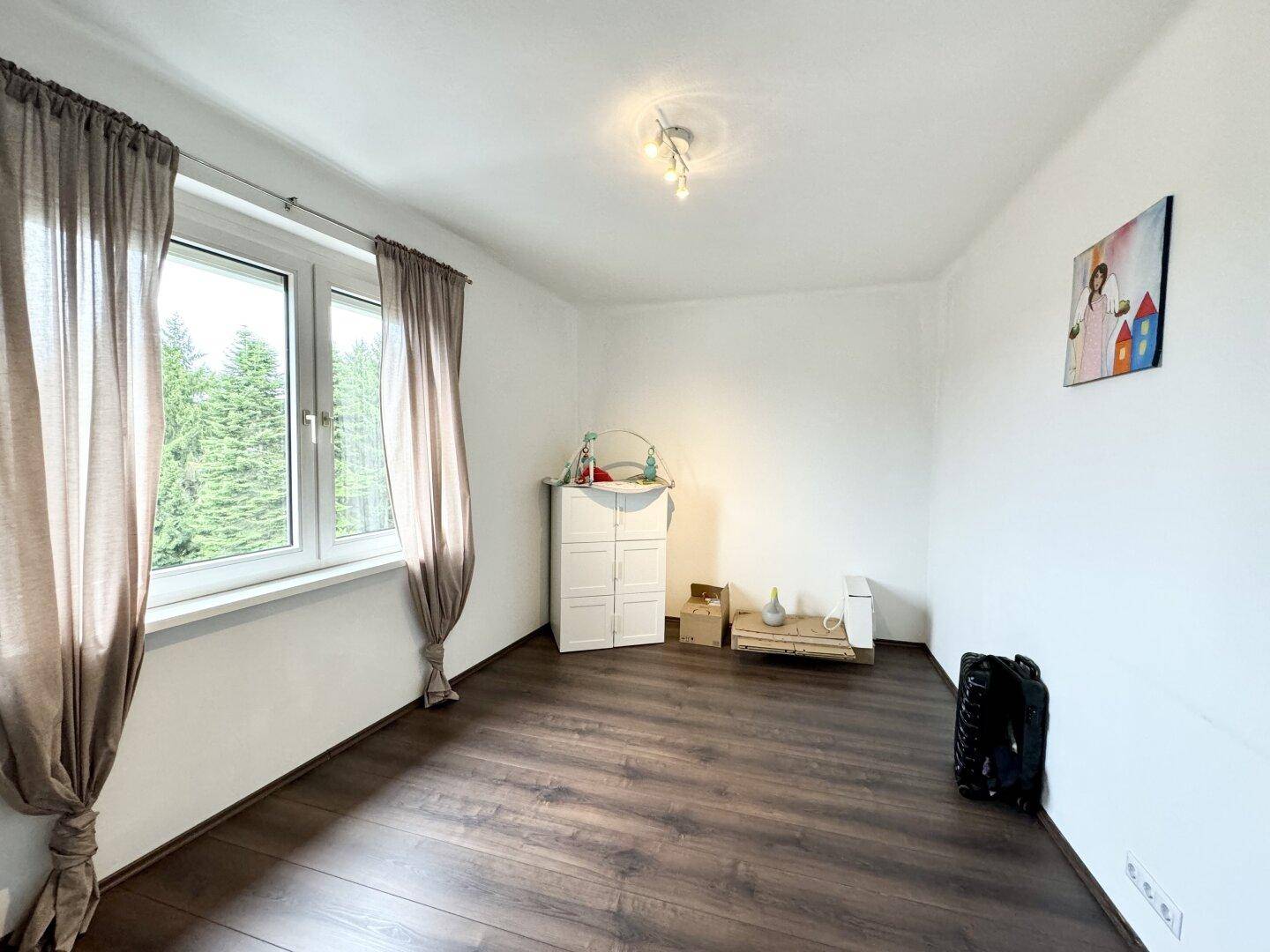 Helles Zimmer mit dunklem Laminatboden und großem Fenster mit Blick ins Grüne.