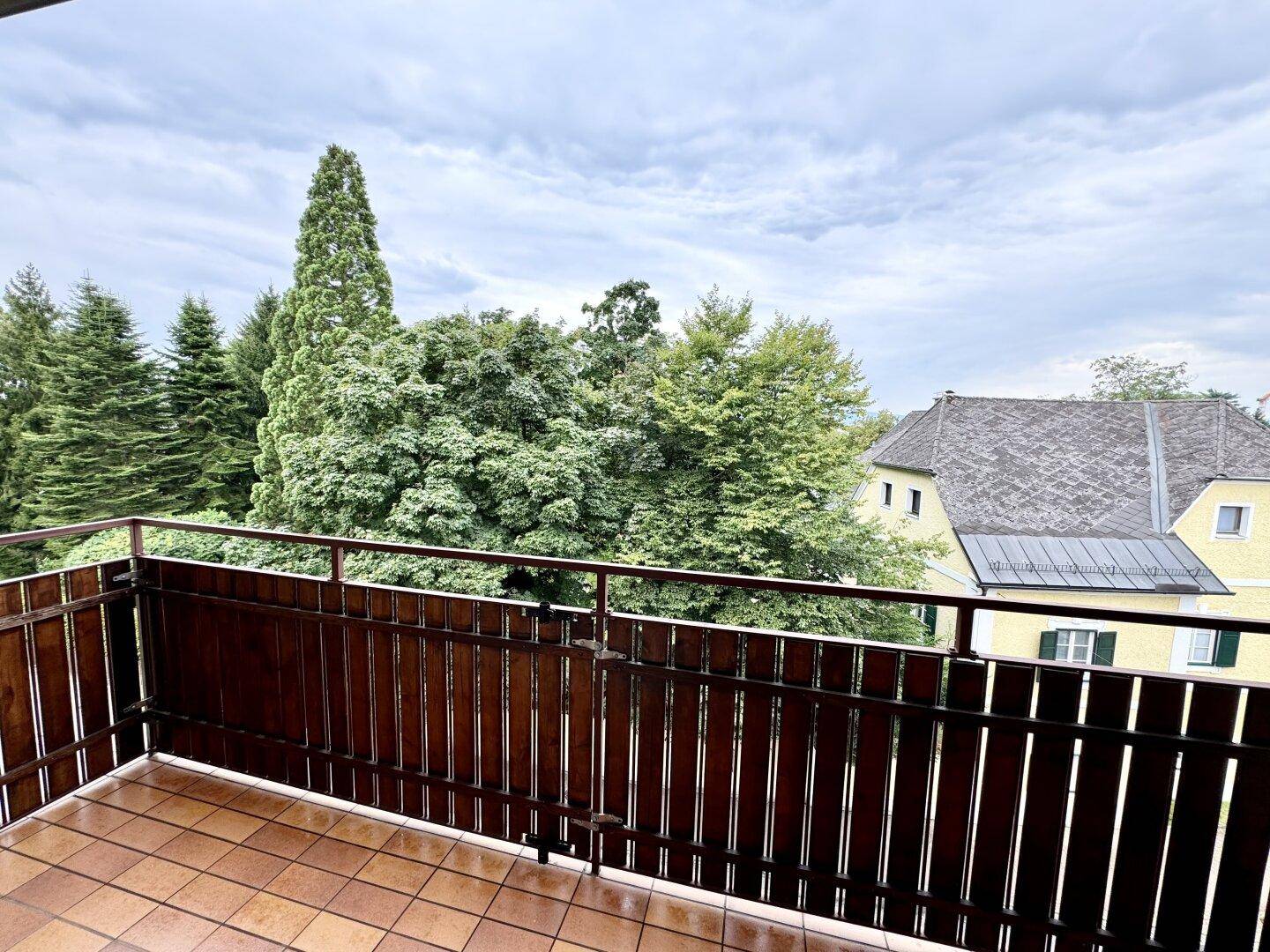 Geräumiger Balkon mit Fliesenboden und Blick auf grüne Bäume und ein charmantes Nachbarhaus.