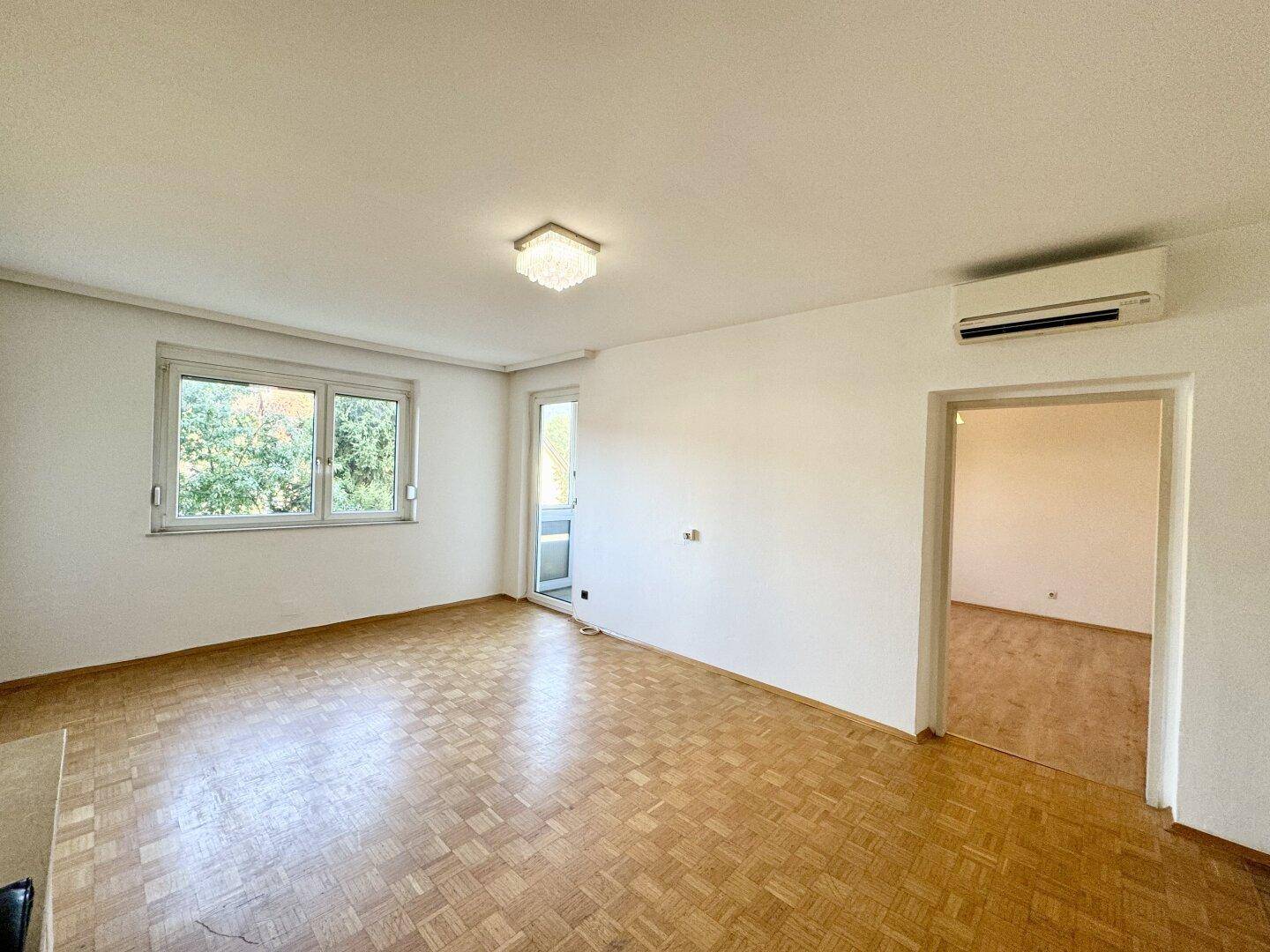 Geräumiges Wohnzimmer mit Parkettboden, großen Fenstern und Zugang zu einem Balkon.