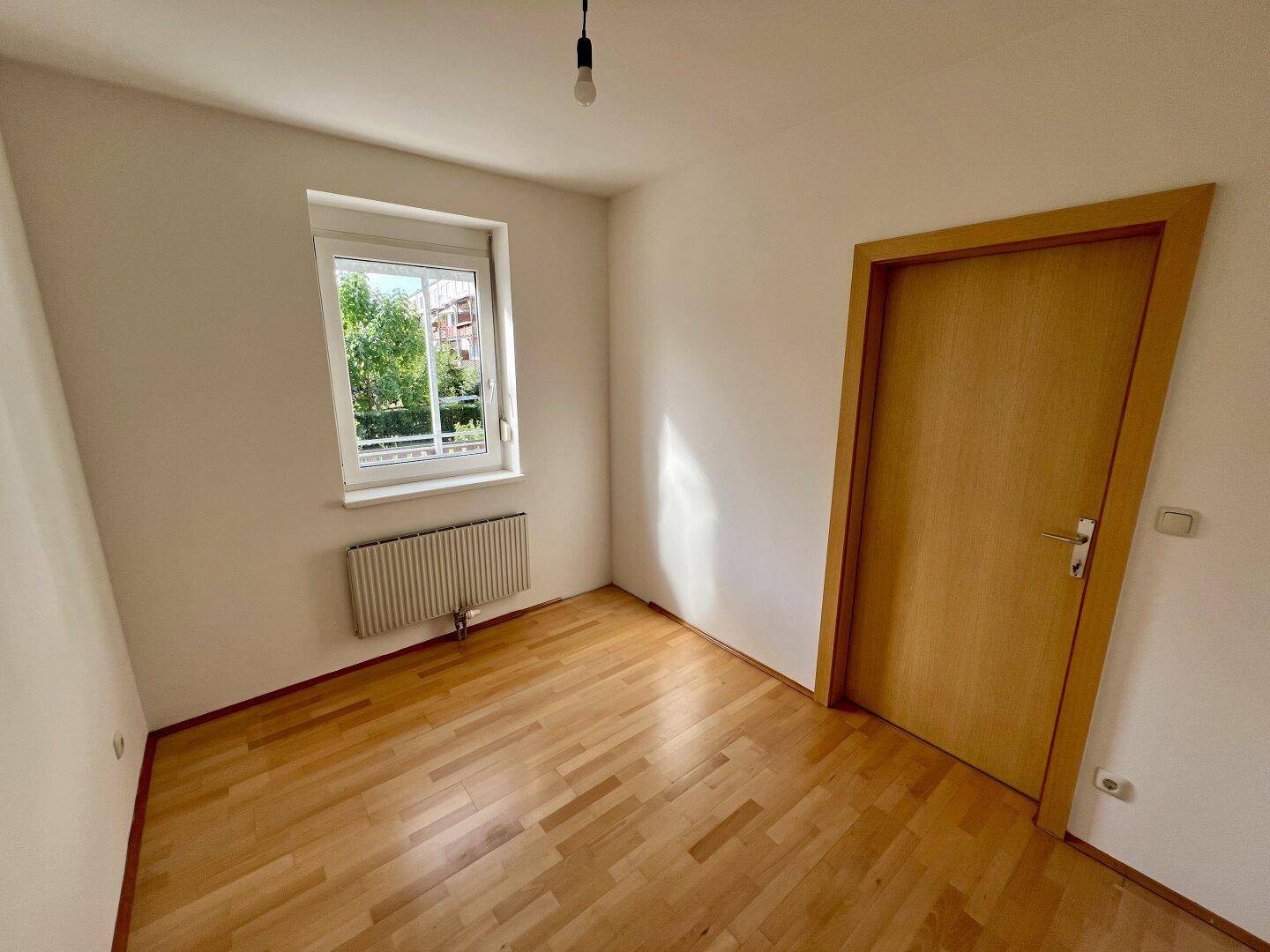 Leeres Schlafzimmer mit hellem Parkettboden, einem Fenster und einem Heizkörper.