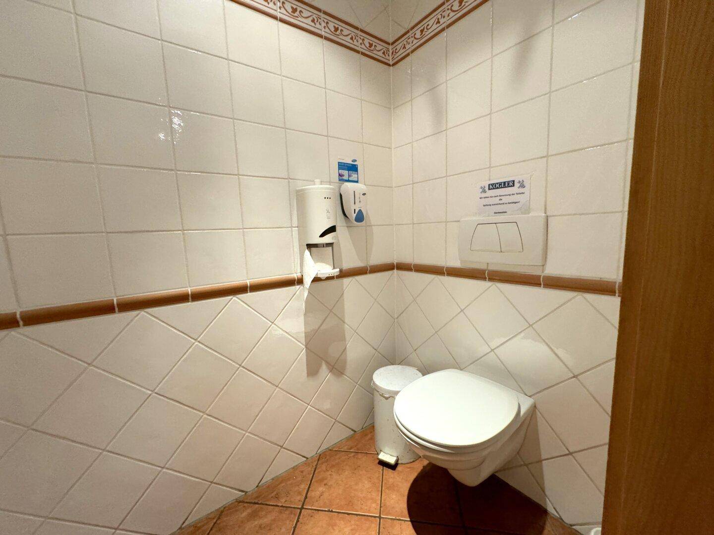Toilettenraum mit weißen Fliesen, Wand-WC und Spender für Desinfektionsmittel.