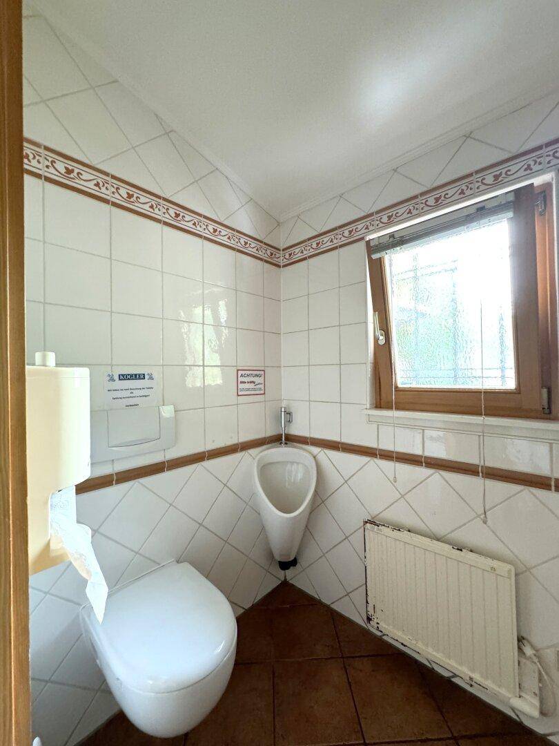 Toilettenraum mit Urinal, Wand-WC, Fenster und Heizkörper.