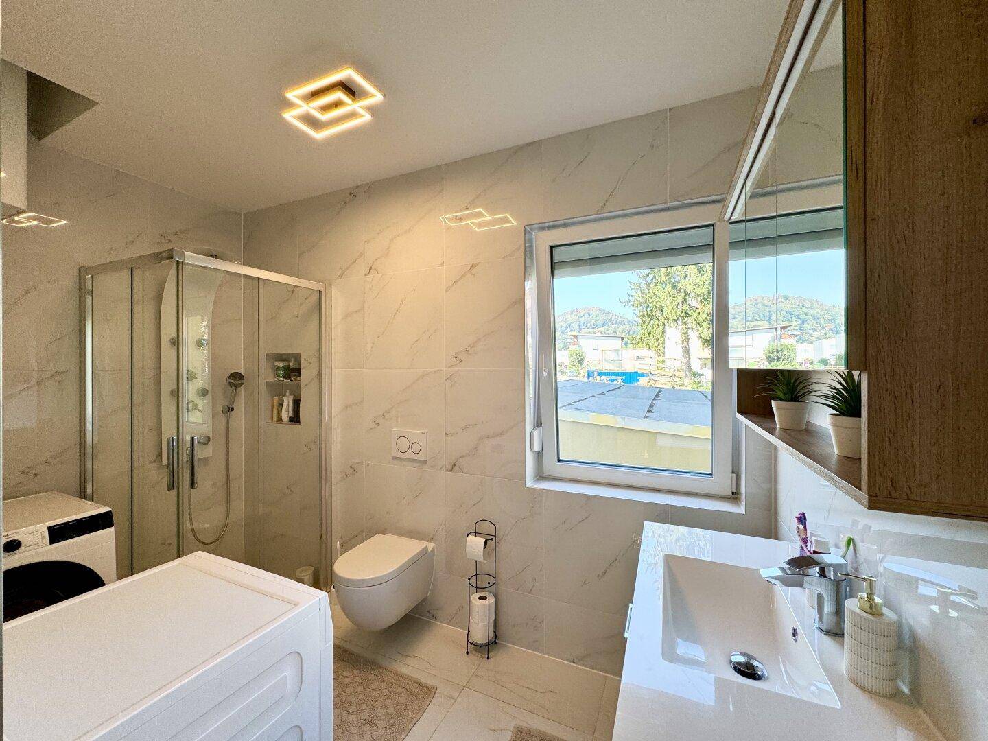 Geräumiges Badezimmer mit Duschkabine, WC, Waschbecken und Waschmaschine, hell gefliest und mit Fenster.