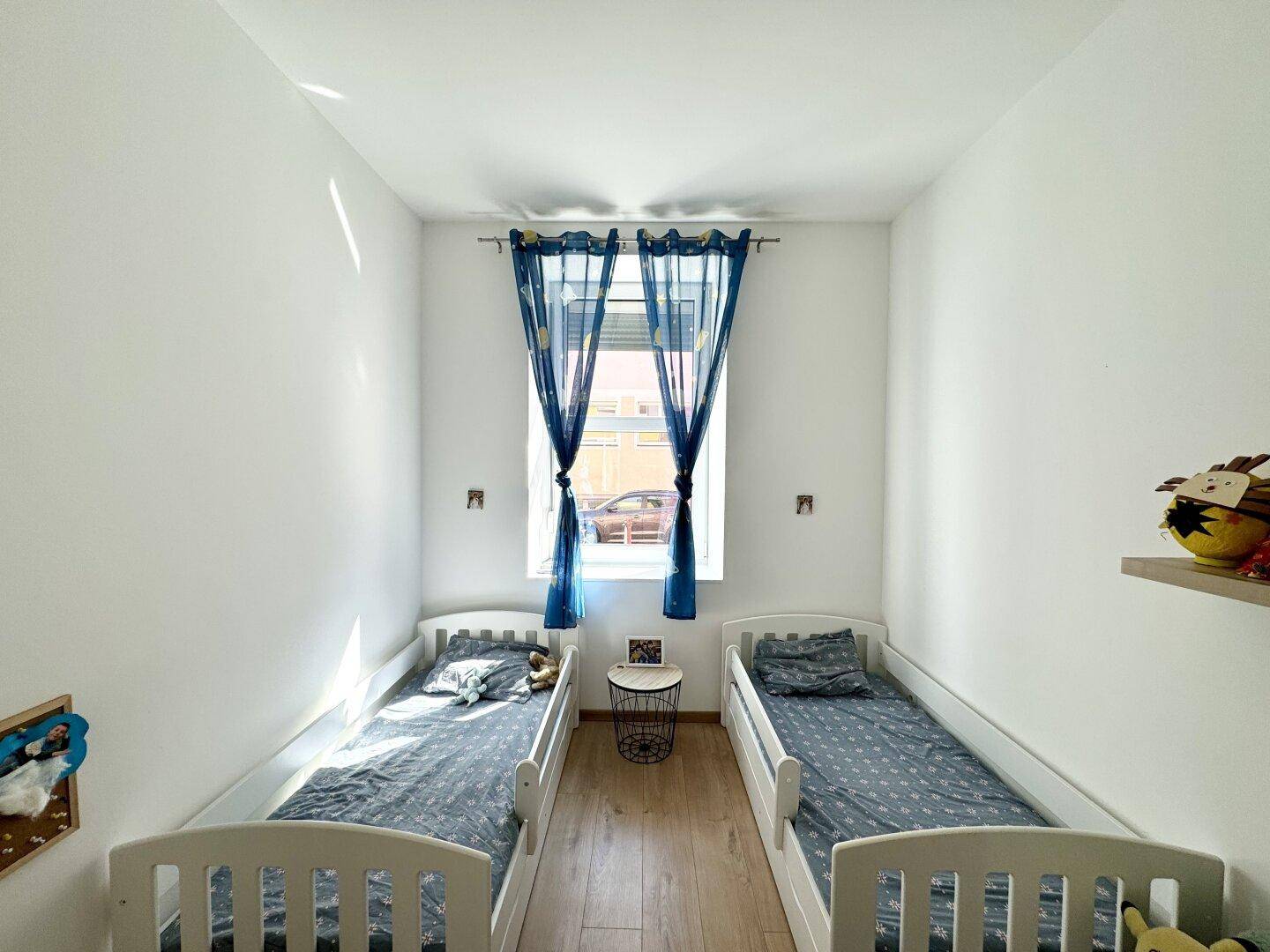 Helles Kinderzimmer mit zwei Einzelbetten und einem Fenster mit blauen Vorhängen, ideal für Geschwister.