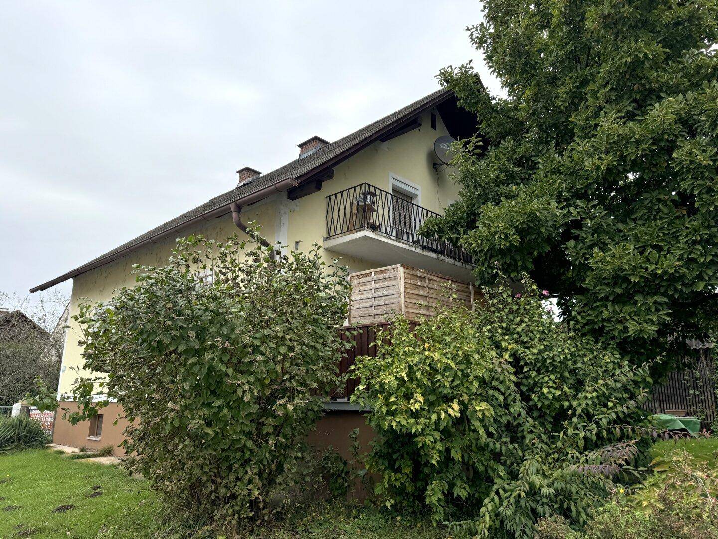 Seitenansicht des Hauses mit kleinem Balkon und umgebenden Bäumen unter bewölktem Himmel.