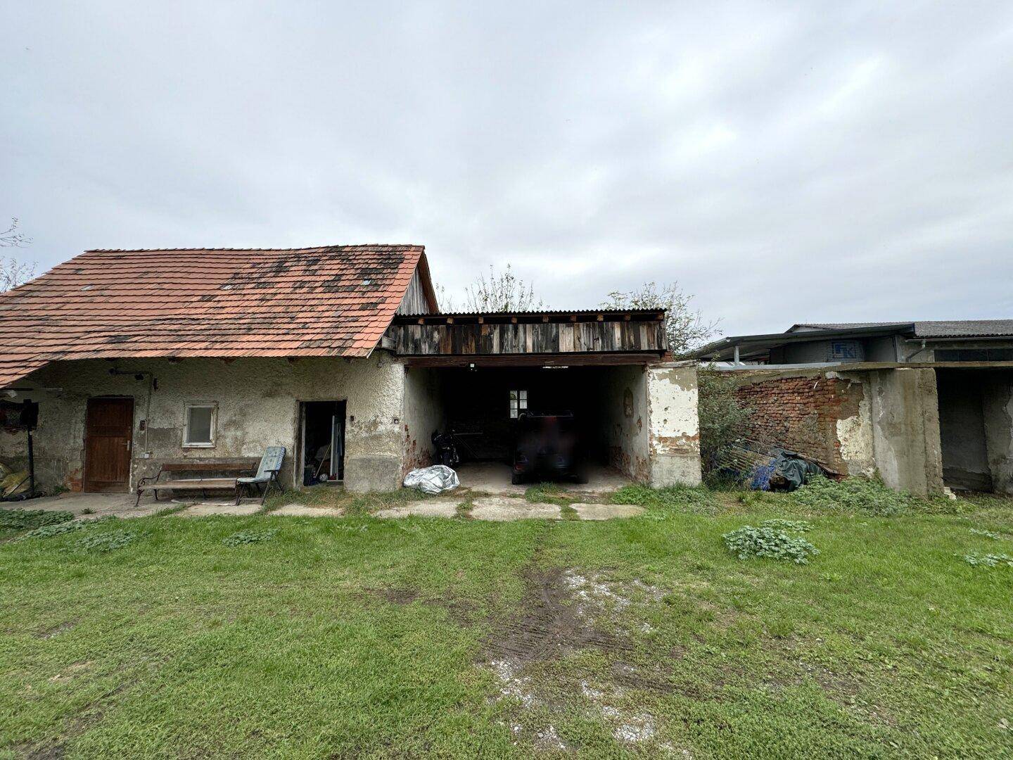 Altes Gebäude mit offenem Garagenbereich und Ziegeldach, umgeben von einer Wiese.