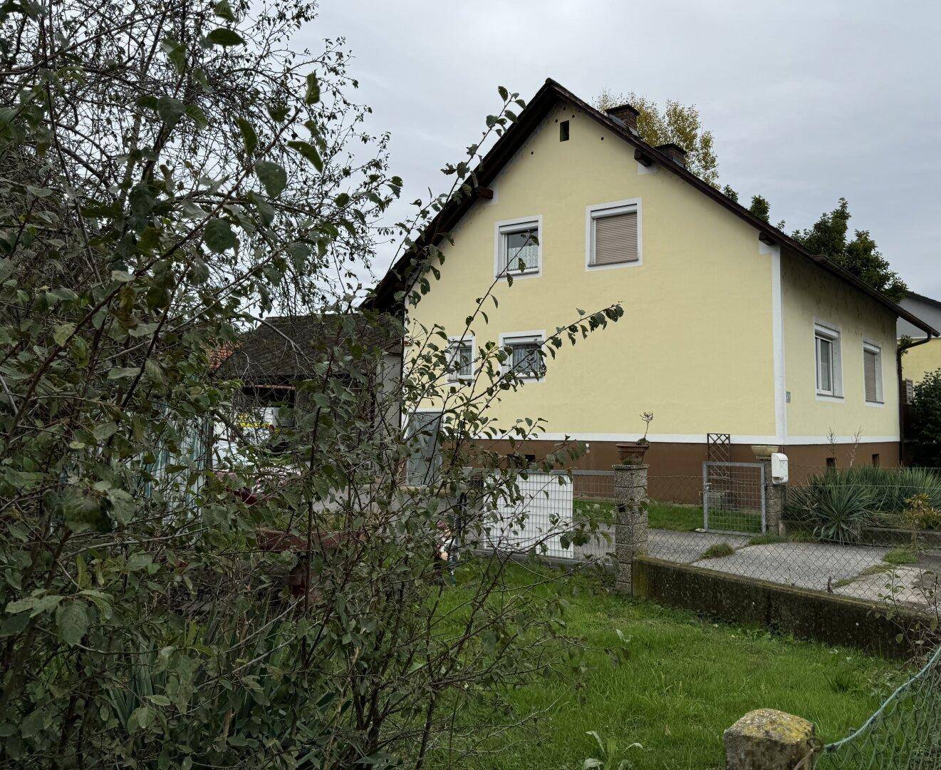 Seitenansicht des gelben Hauses, teilweise von Bäumen verdeckt, unter bewölktem Himmel.