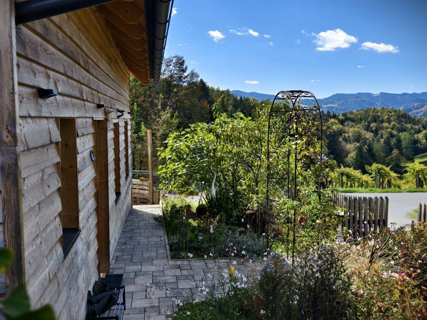 Seitenansicht des Hauses mit gepflastertem Weg, blühendem Garten und Bergblick.