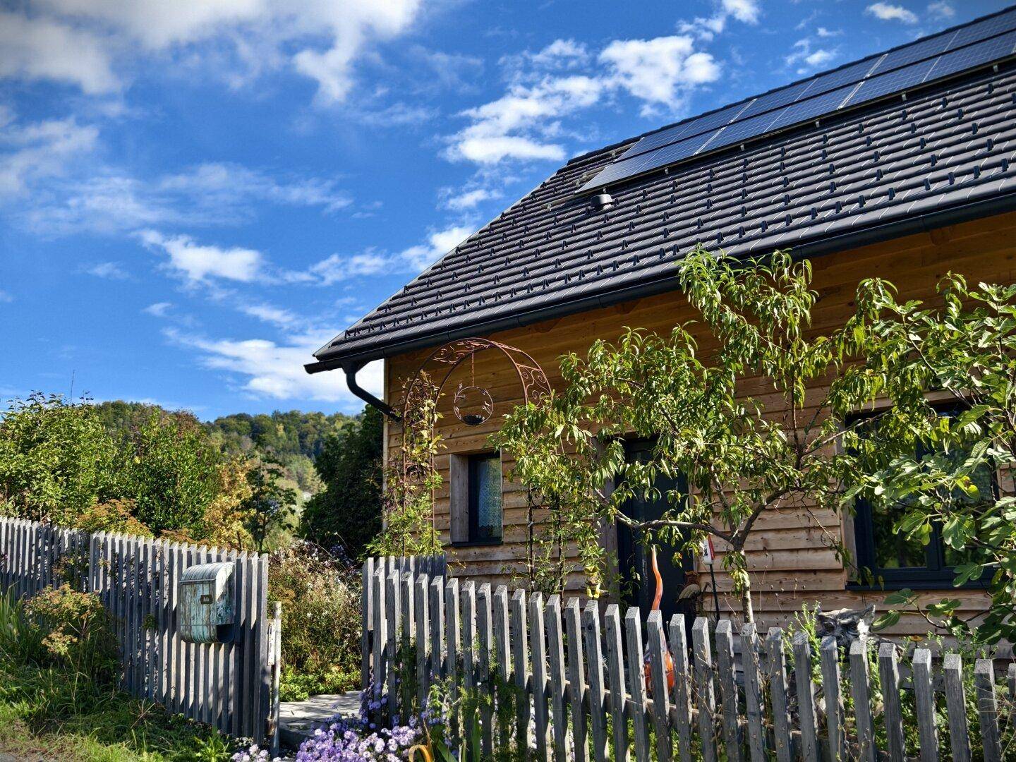 Vorderansicht des Hauses mit Holzzaun, Garten und Solarpaneelen auf dem Dach.