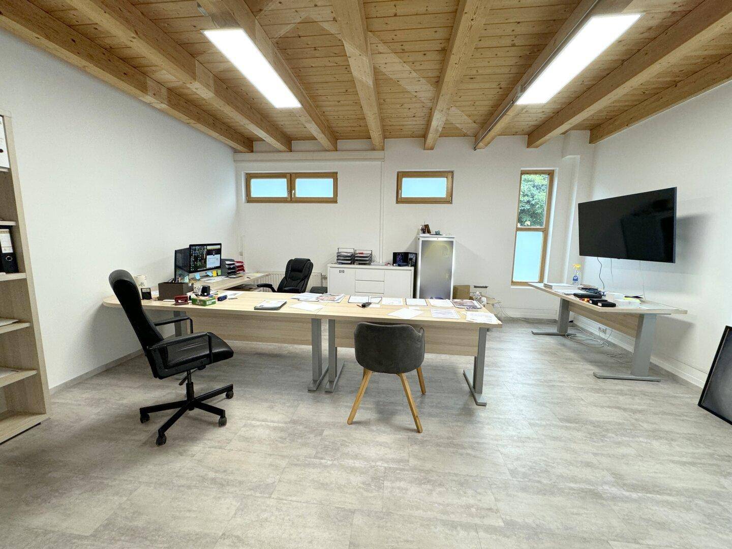 Helles Büro mit Holzbalkendecke, mehreren Arbeitsplätzen und großen Fenstern, die viel Tageslicht hereinlassen.
