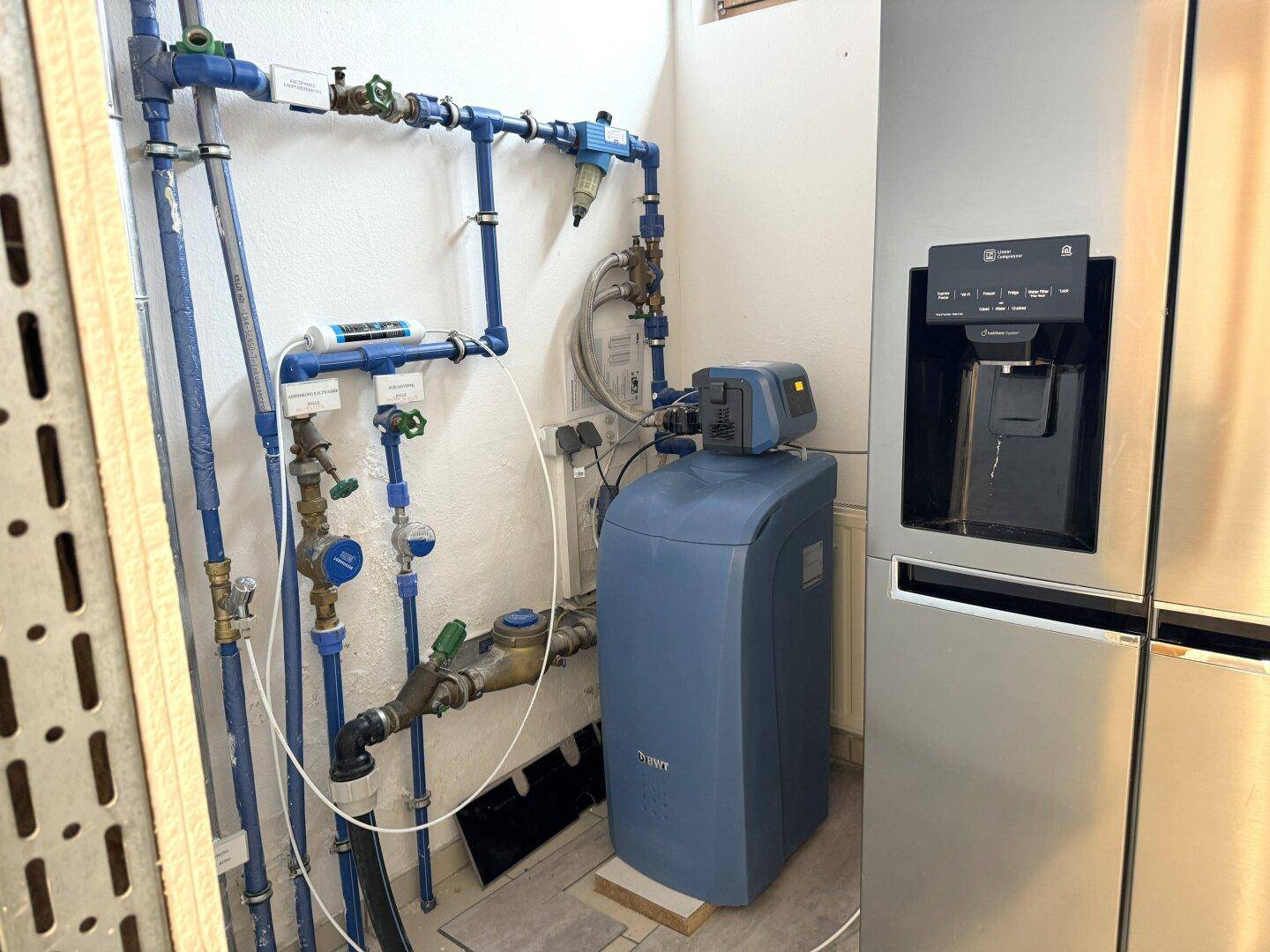 Technikraum mit sichtbaren blauen Wasserleitungen, einem Warmwasserspeicher und einem großen Kühlschrank.