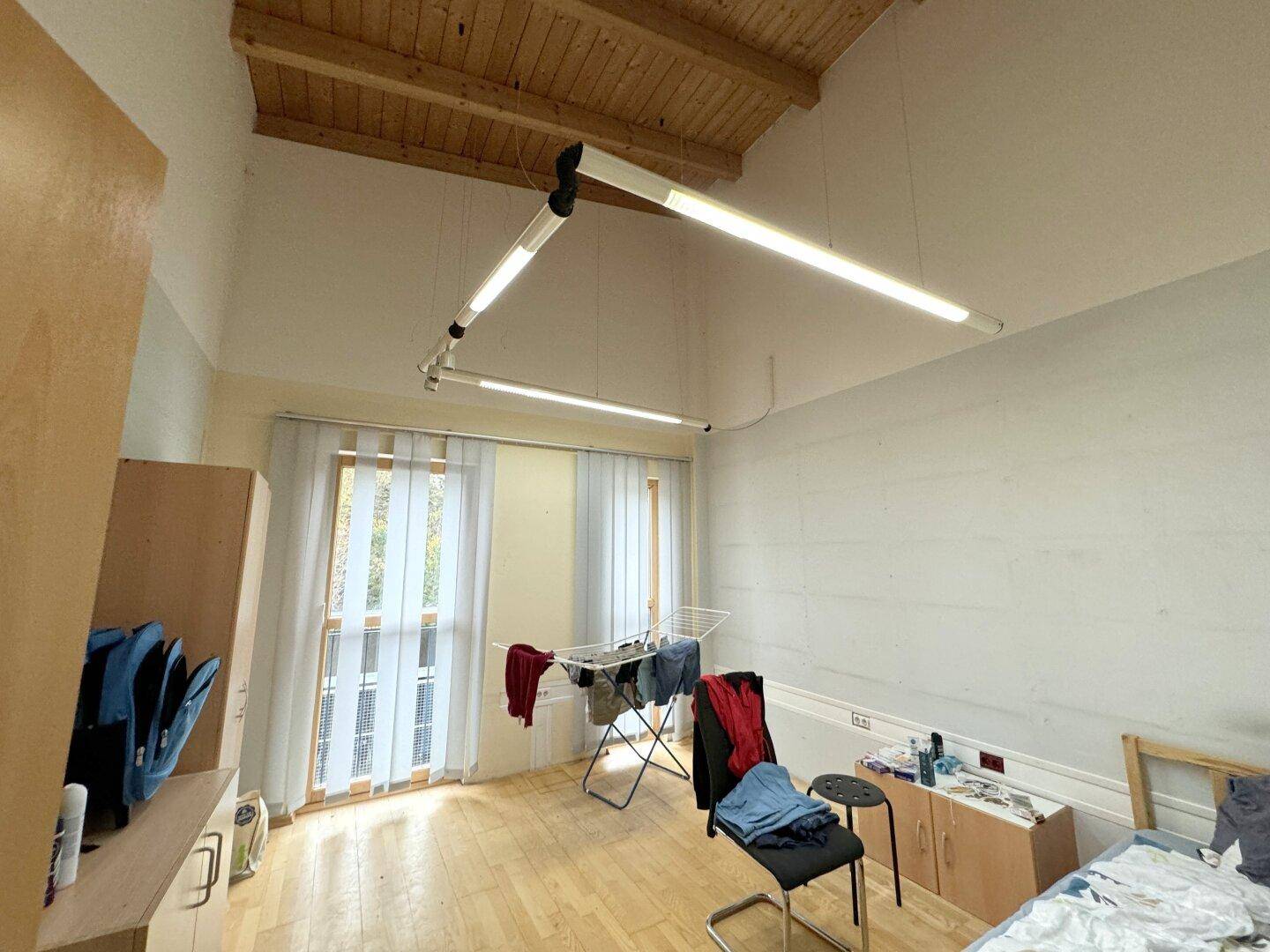 Schlafzimmer mit Holzbalkendecke, Holzfußboden und großen Fenstern, die viel Licht hereinlassen.