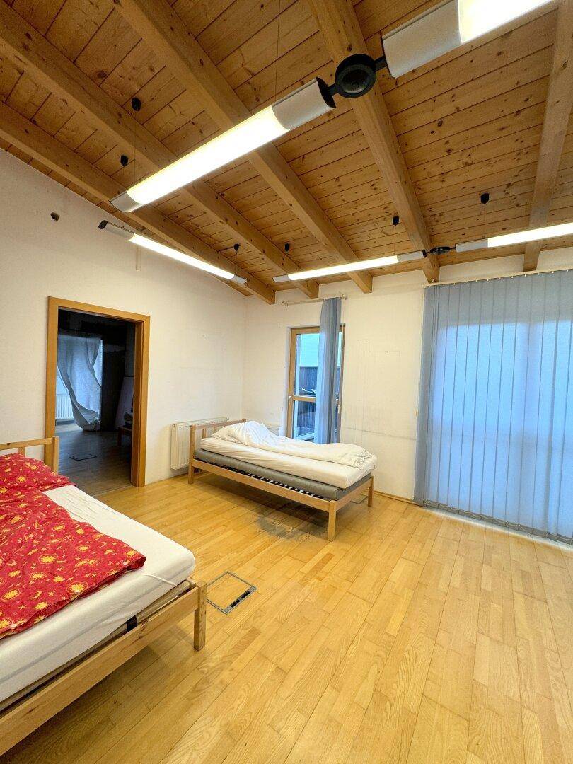 Schlafzimmer mit Holzbalkendecke, Holzfußboden und einem großen Fenster mit Jalousien.