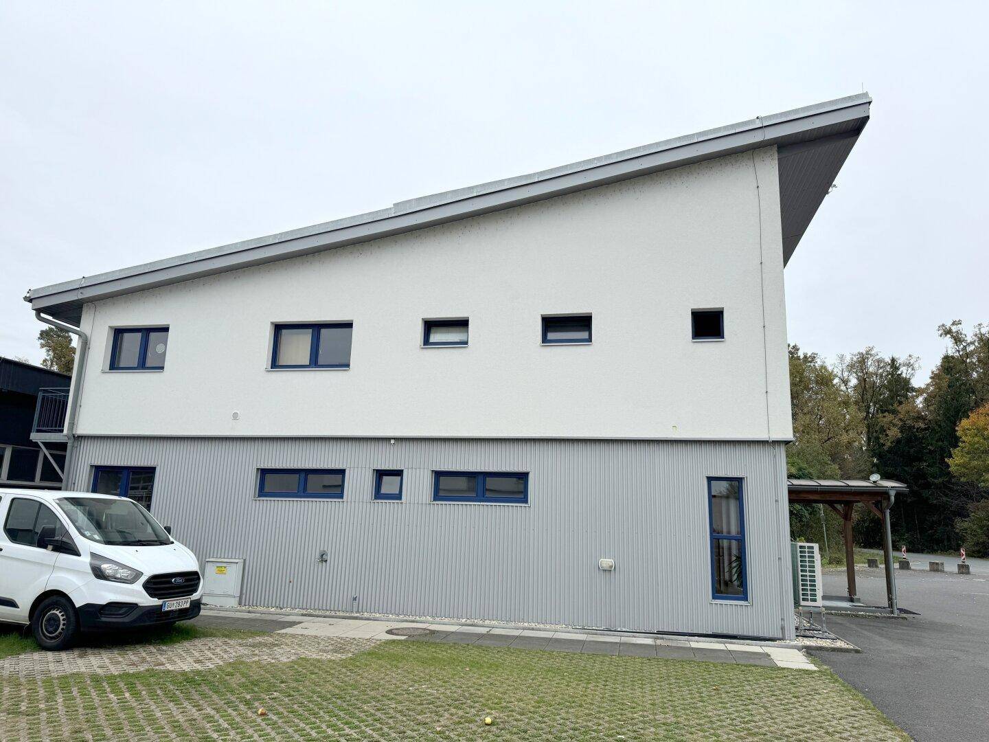 Modernes Gebäude mit schrägem Dach, weißer Fassade und blauen Fenstern, umgeben von Grünflächen.