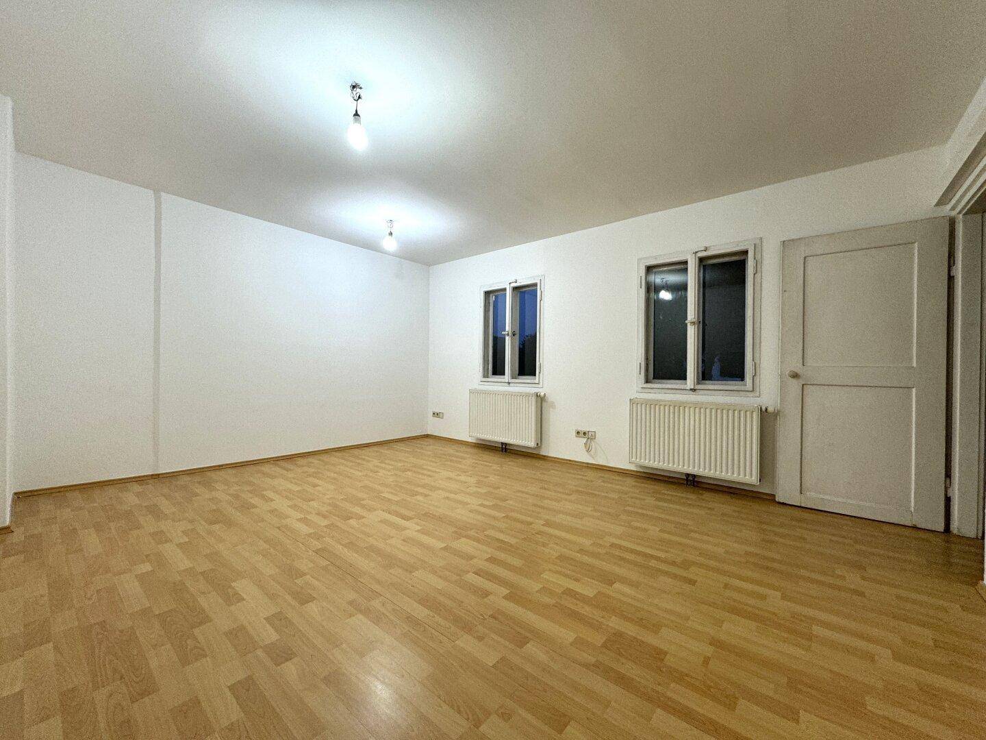 Heller, leerer Raum mit Laminatboden, weißen Wänden und zwei Fenstern.