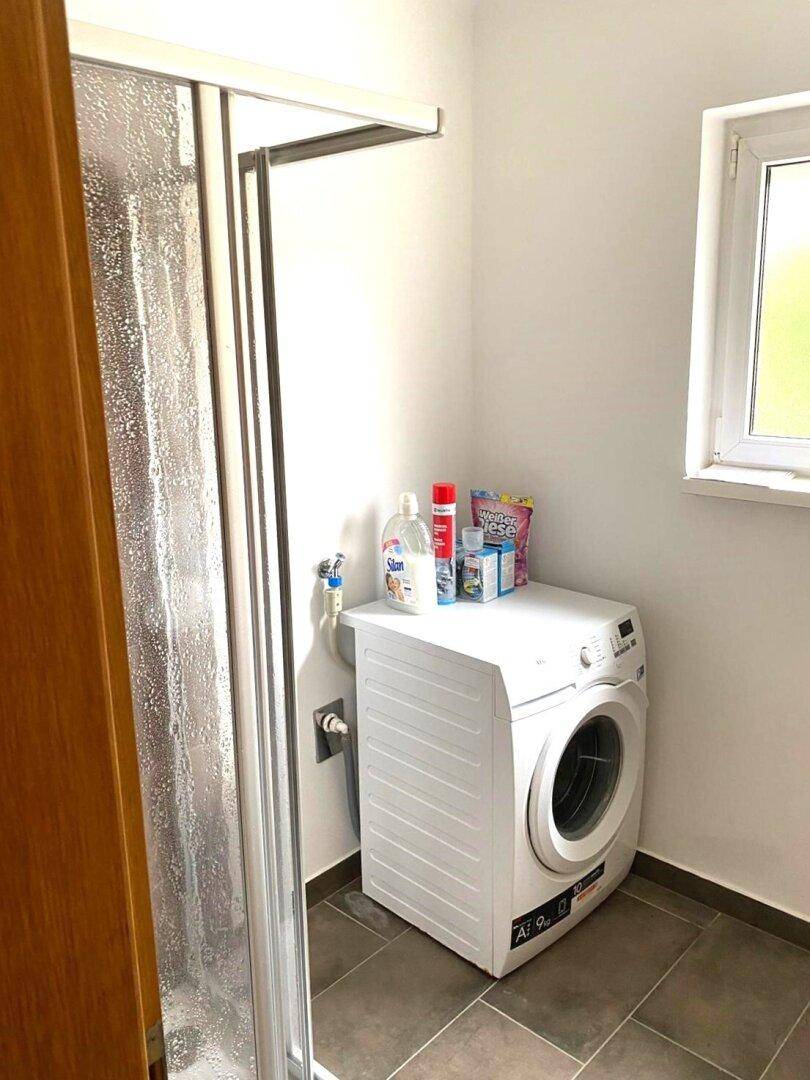 Kleiner Waschraum mit Waschmaschine, Duschkabine und Fenster für Belüftung.